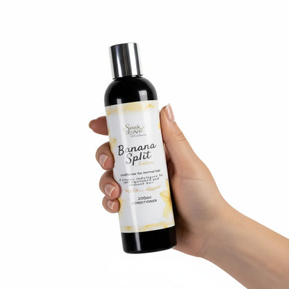 Hand holding a 200ml bottle of Soak in Love Banana Split Edition Conditioner for normal hair with a white and yellow label against a white background.